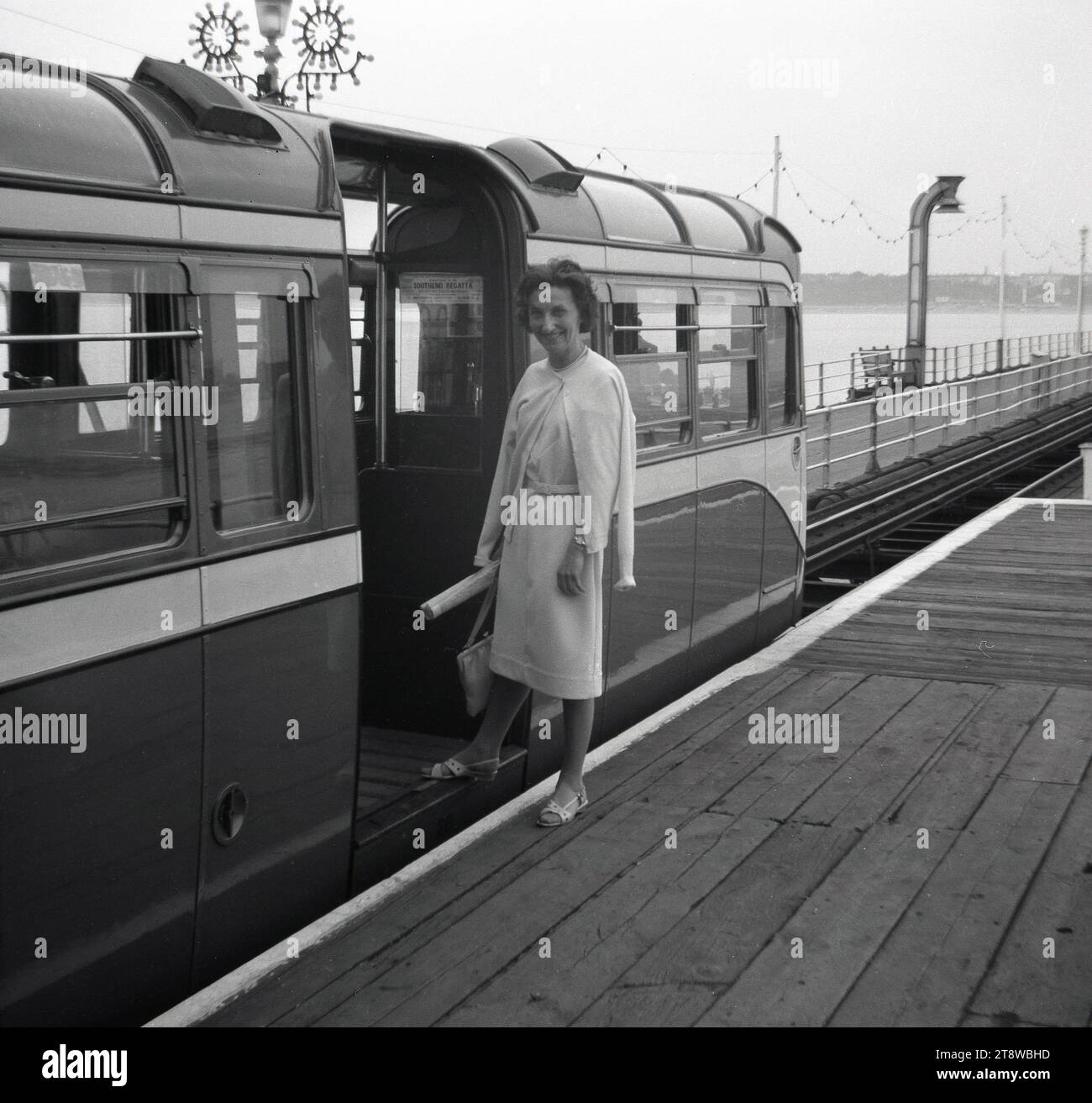 Anni '1950, storica, una donna ben vestita in piedi su una piattaforma ferroviaria di legno per salire a bordo di un treno elettrico al molo di Southend on Sea. All'interno della carrozza ferroviaria, un avviso per la Southend Regatta. Questi vagoni ferroviari del molo furono introdotti nel 1949 e funzionarono fino alla fine degli anni '1970, quando fu aperto un servizio ferroviario diesel più piccolo. Foto Stock