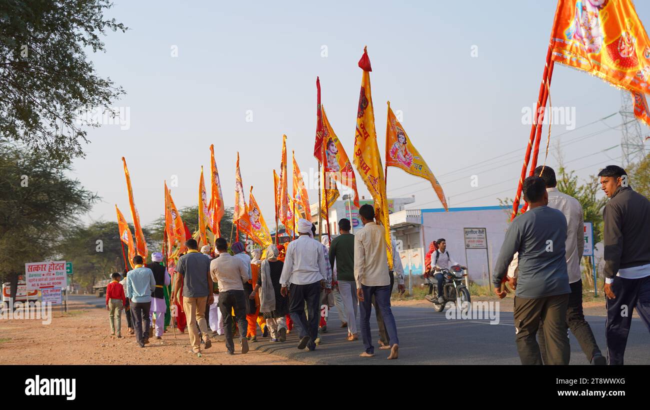 15 novembre 2023 Khatushyamji, Rajasthan, India. Persone che trasportano Kalash di rame con bandiere su bastoni. Bandiere fluttuanti gialle con colori brillanti riga di bandiere Foto Stock