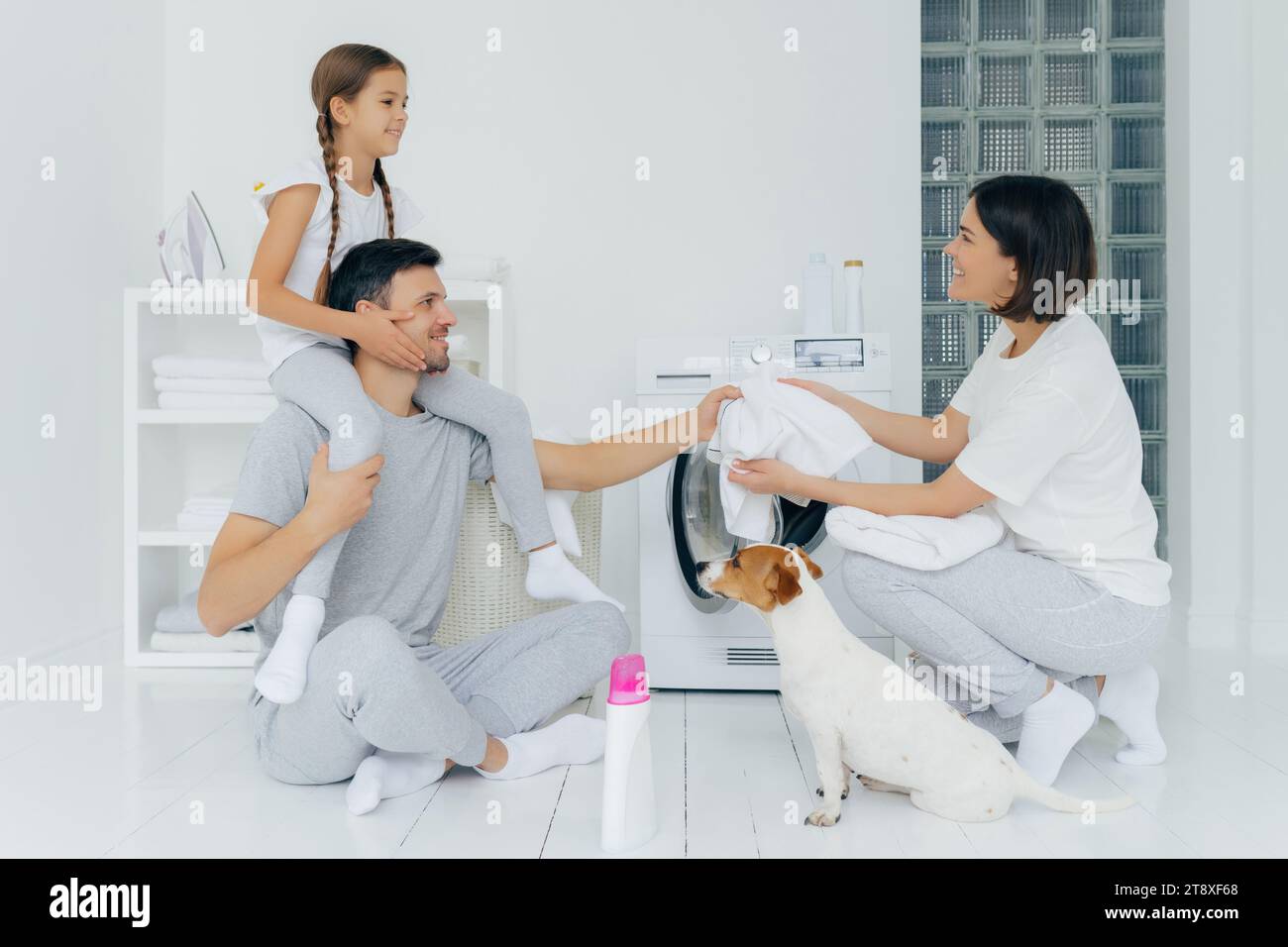 Famiglia con un bambino e un cane che si lavano insieme in una lavanderia bianca e organizzata Foto Stock