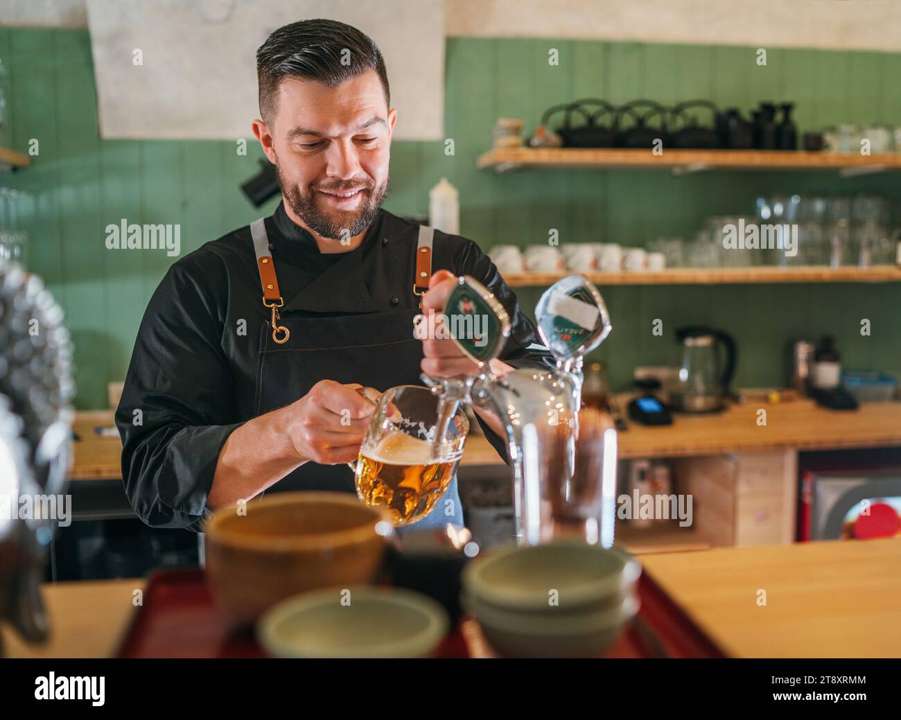Barman barbarbarocco sorridente vestito di uniforme nera con un grembiule che sbatte birra fresca lager in una tazza di vetro al bancone del bar. Persone di successo, truffa della birra Foto Stock