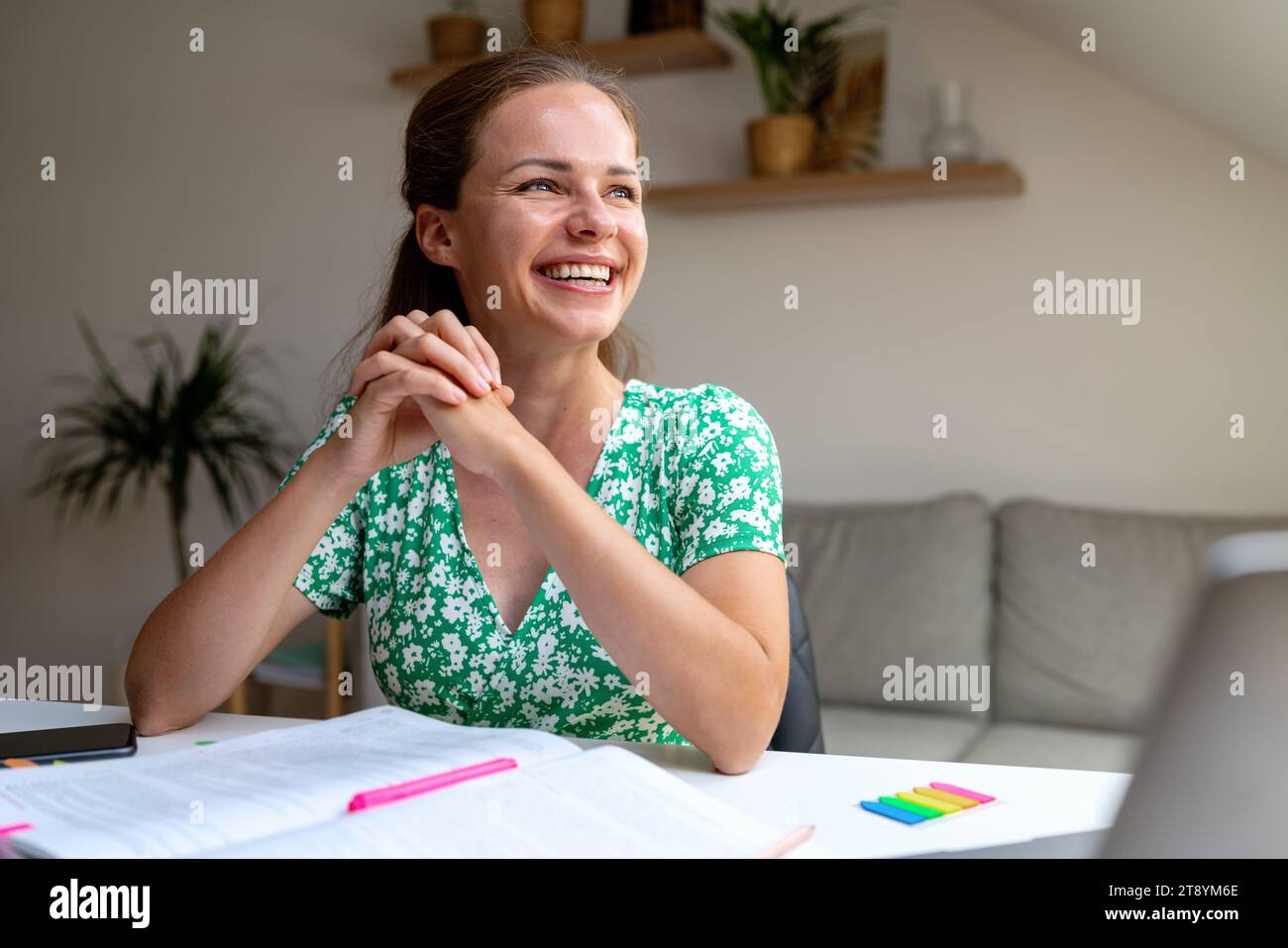 Studentessa adulta ispirata e motivata ride mentre fa i compiti a casa seduta alla scrivania. Foto Stock