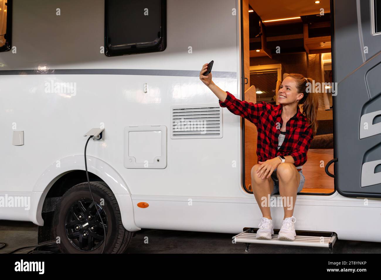 Giovane donna che si autoritratta mentre si trova seduto sulla porta del camper per per veicoli ricreativi. Stile di vita del parcheggio per camper. Foto Stock