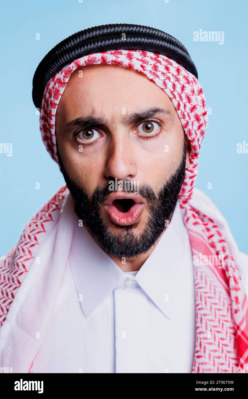 Uomo musulmano con bocca aperta che indossa il tradizionale foulard ghutra a scacchi ritratto da studio per primo piano. Persona araba con copricapo islamico che mostra emozioni sul viso e guarda la fotocamera Foto Stock