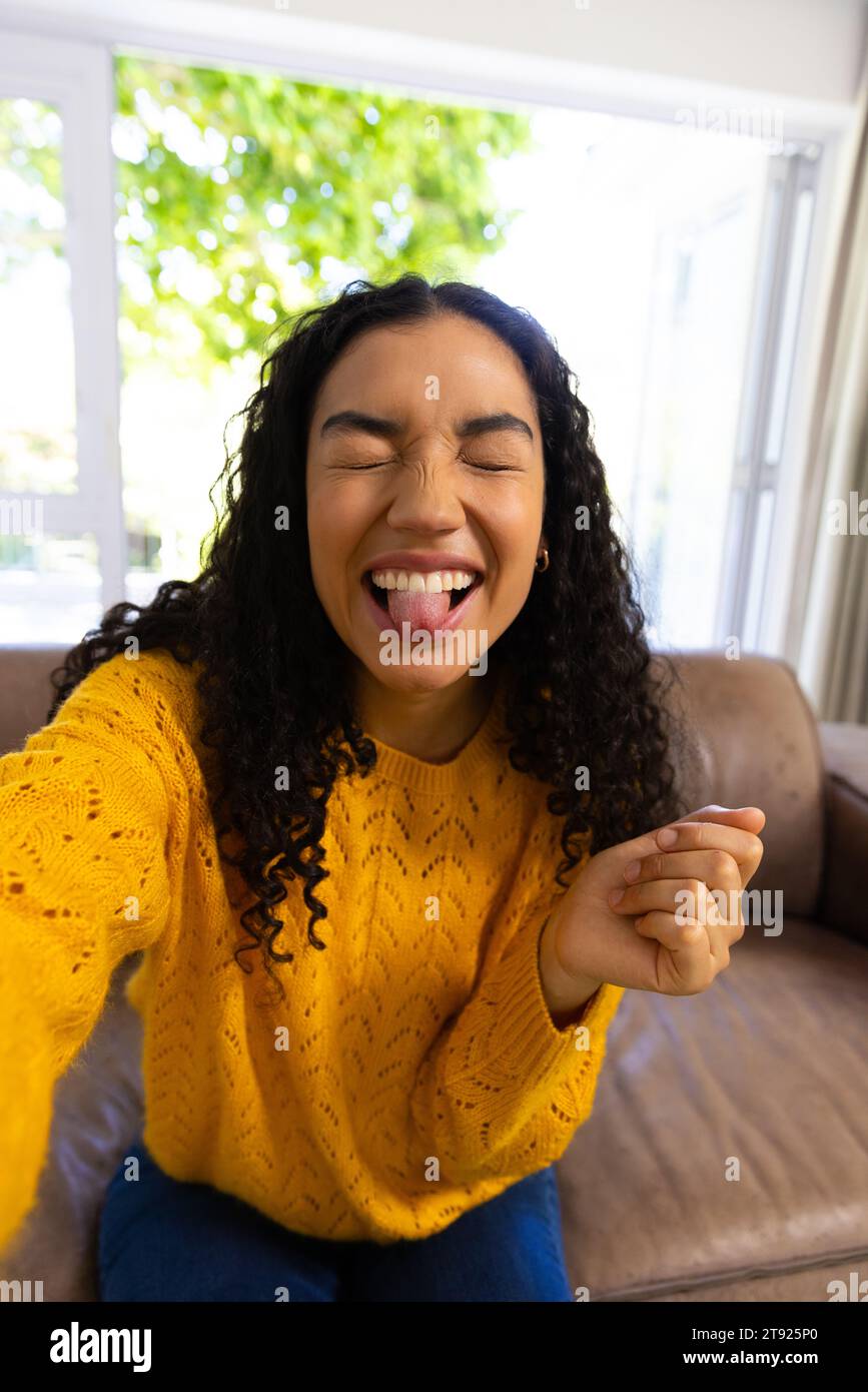 Felice donna birazziale con maglione giallo che effettua videochiamate e si prende gioco a casa Foto Stock