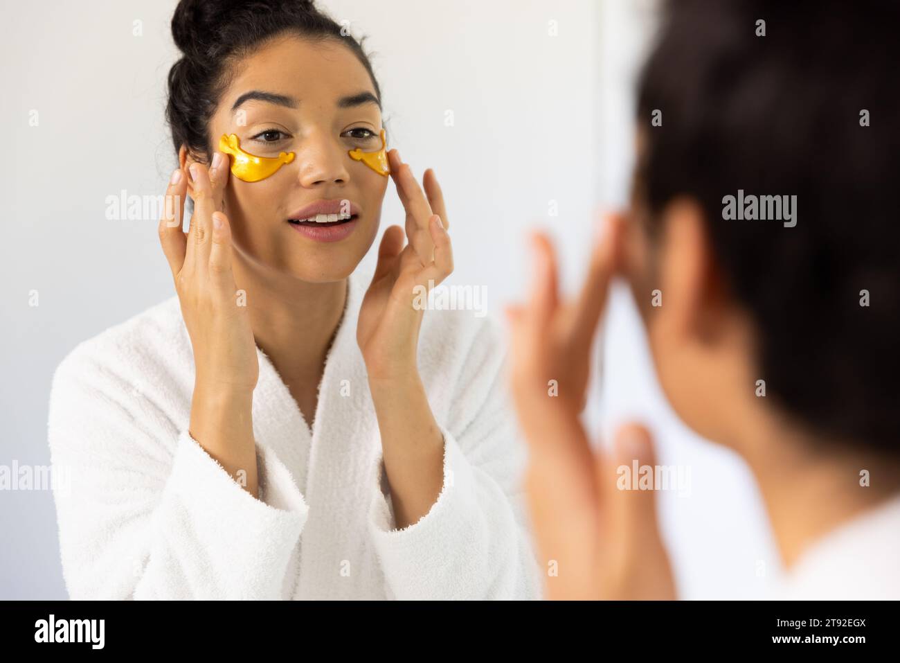 Donna birazziale in accappatoio che applica una maschera per gli occhi nel bagno soleggiato. Stile di vita, cura di sé, bellezza, pelle e vita domestica, inalterati. Foto Stock