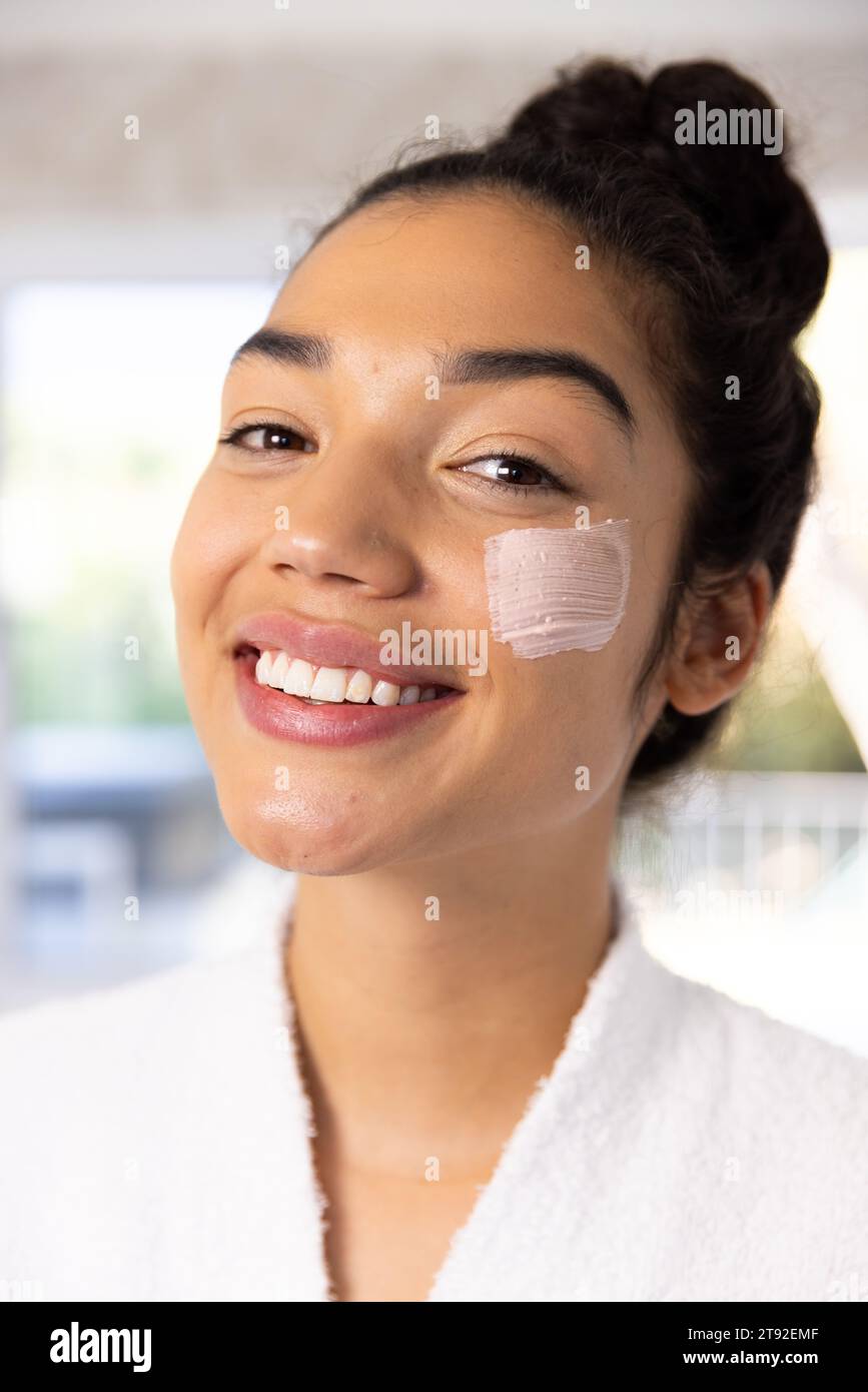 Ritratto di felice donna birazziale in accappatoio applicazione maschera viso in bagno soleggiato. Stile di vita, cura di sé, bellezza, pelle e vita domestica, inalterati. Foto Stock