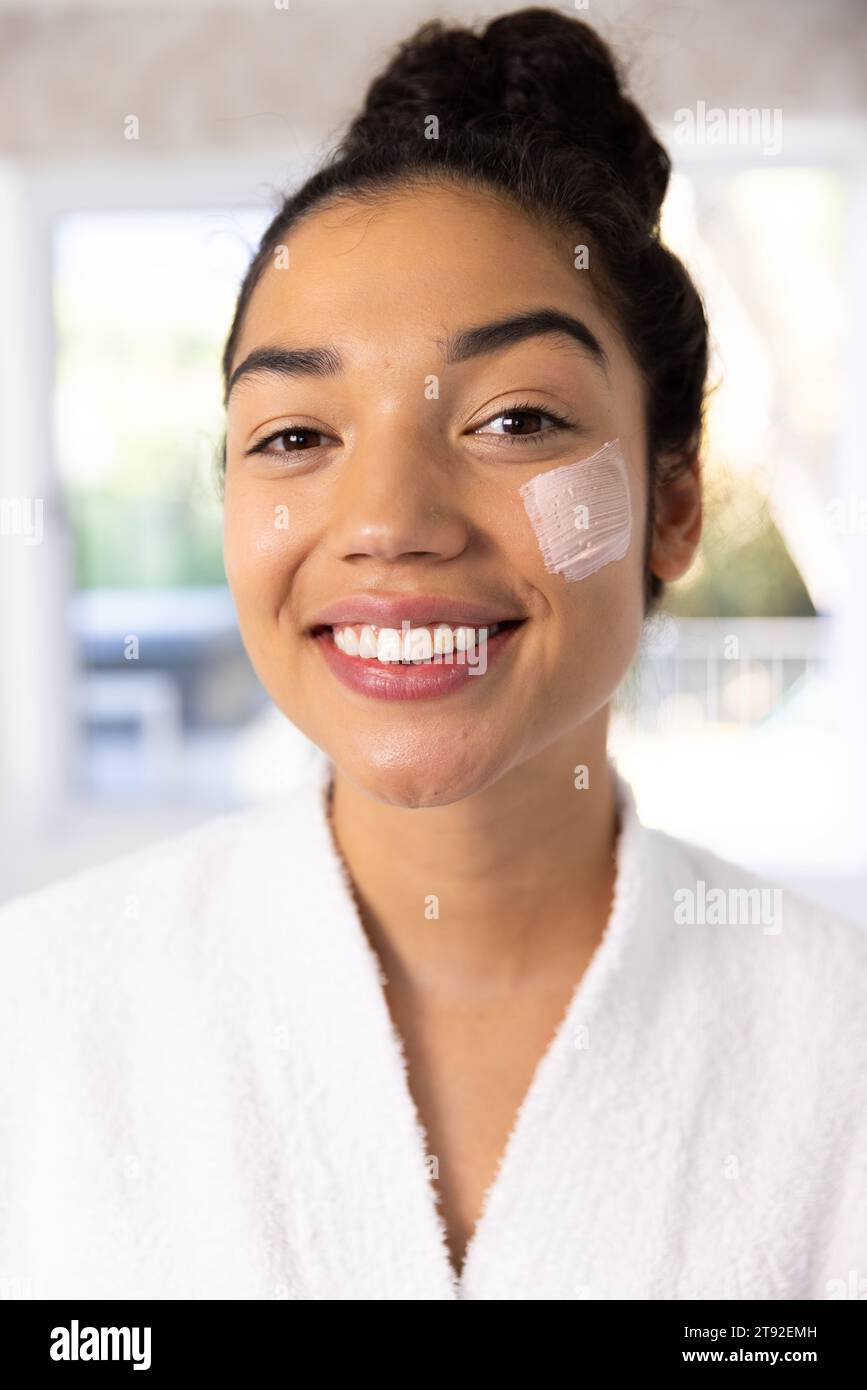 Ritratto di felice donna birazziale in accappatoio applicazione maschera viso in bagno soleggiato. Stile di vita, cura di sé, bellezza, pelle e vita domestica, inalterati. Foto Stock