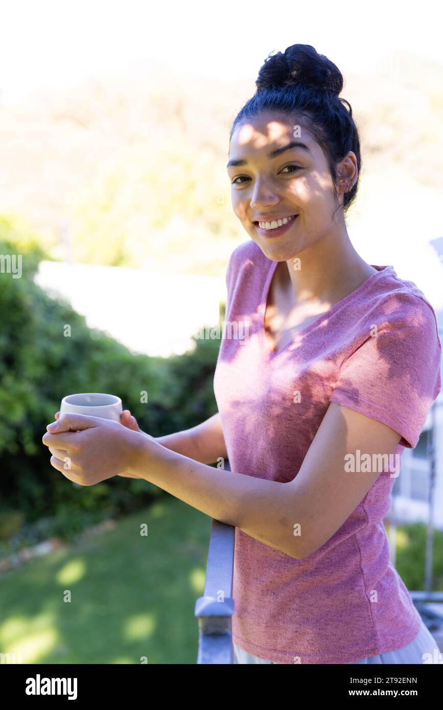 Ritratto di felice donna birazziale che tiene una tazza di caffè sulla terrazza soleggiata. Stile di vita, tempo libero, natura e vita domestica, inalterati. Foto Stock