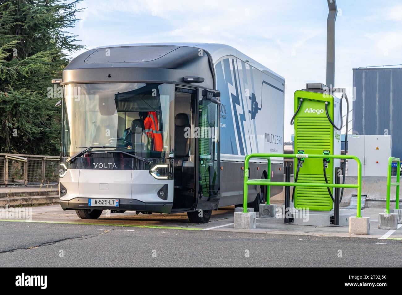 Un camion volta Zero, porta aperta, è parcheggiato in una stazione di ricarica Allego, con alberi a sinistra e un cielo blu sullo sfondo. Foto Stock