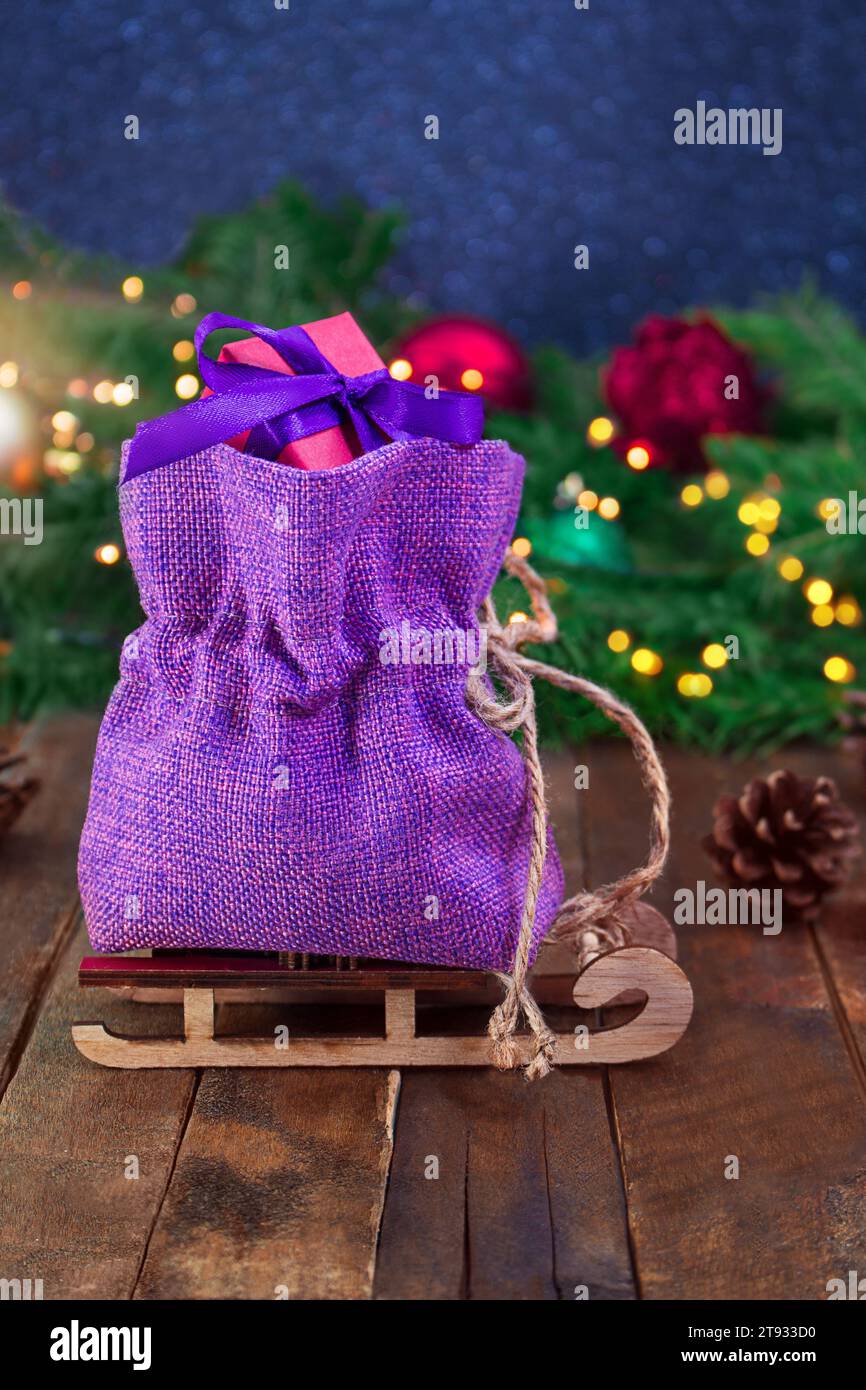 Borsa in tela viola con confezione regalo rossa su slitta su tavole di legno. Rami di abete rosso, giocattoli e luci sfocate. Natale, Capodanno, saldi. Messa a fuoco morbida. Eveni Foto Stock