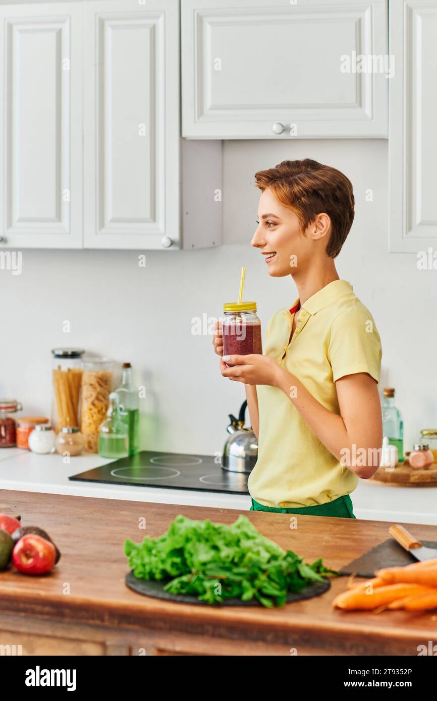 vista laterale di una donna vegetariana sorridente che tiene il barattolo mason con frullato fresco in cucina moderna Foto Stock