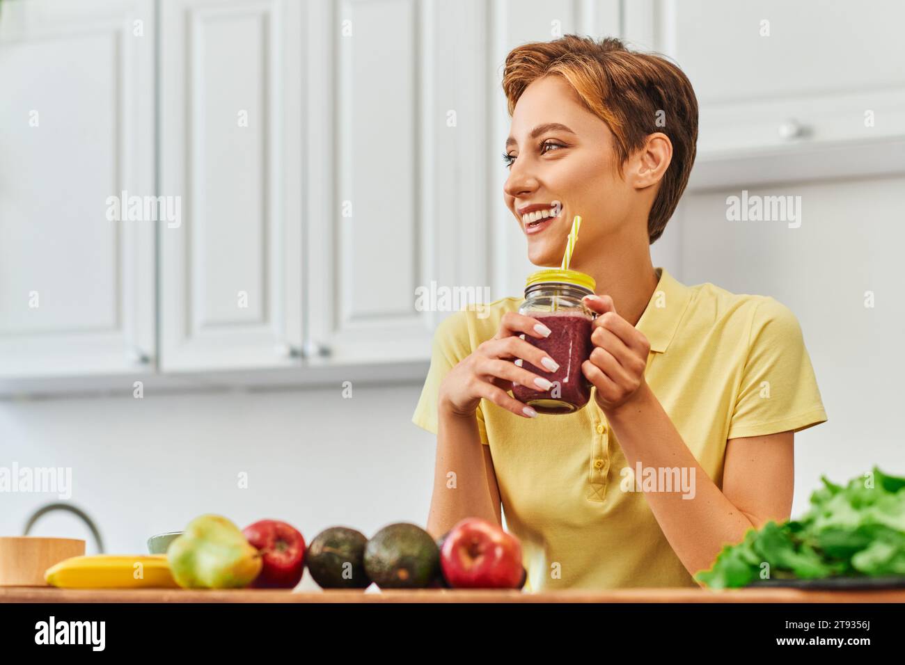 donna vegetariana allegra che tiene frullato fresco in barattolo mason con paglia e guarda lontano in cucina Foto Stock