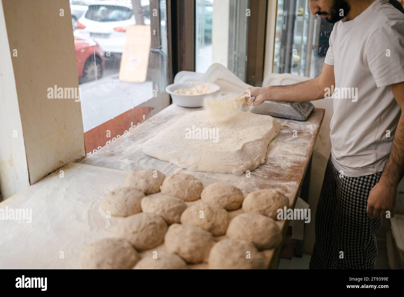 Giovane panettiere arabo con t-shirt bianca versando farina sull'impasto con panini di pasta accanto ad esso su un tavolo in una panetteria Foto Stock