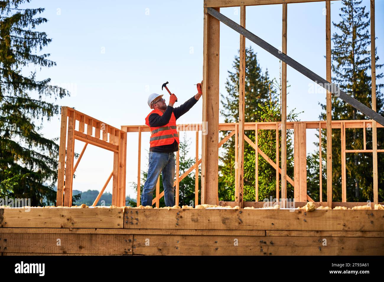 La casa costruita con cornici in legno è stata eretta da falegname vicino alla foresta. Uomo barbuto che indossa occhiali che guida con le unghie con il martello mentre indossa un casco di sicurezza e un gilet da costruzione. Foto Stock