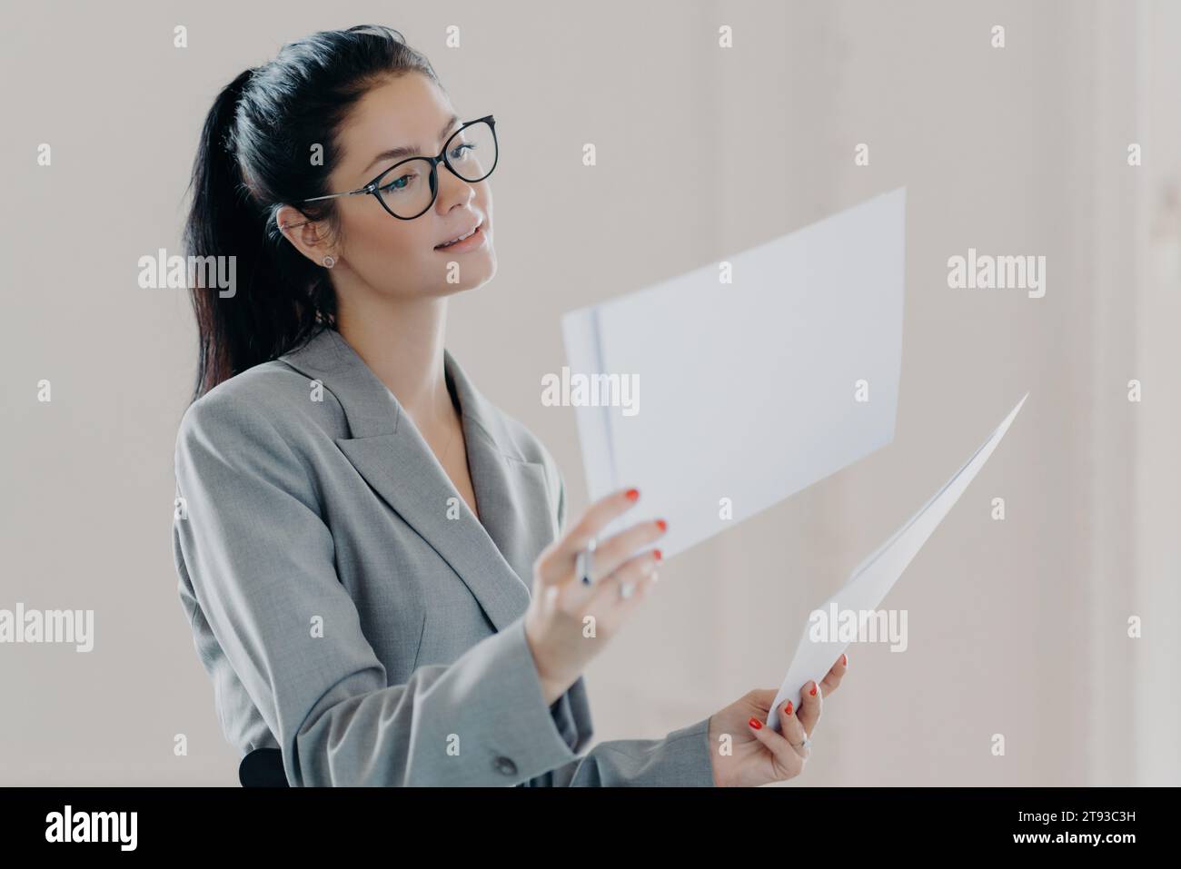 Donna d'affari attenta che esamina i documenti, indossa occhiali e un abito grigio su uno sfondo chiaro. Foto Stock