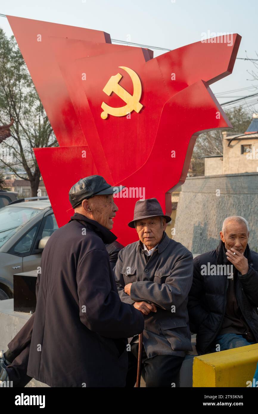 Gli anziani siedono di fronte a una scultura di una bandiera rossa con il logo del Partito Comunista a Pechino, in Cina. 21 novembre 2023 Foto Stock