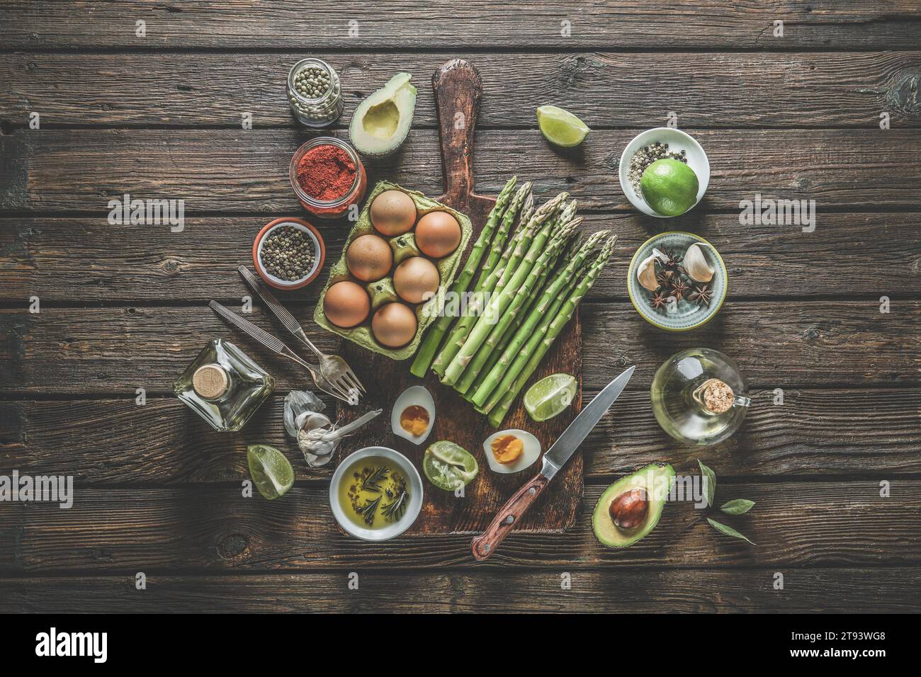 Vari ingredienti sani di cottura: Mazzo di asparagi verdi, uova, spezie, avocado, lime, olio d'oliva e utensili da cucina: forchette, coltelli e taglio b Foto Stock
