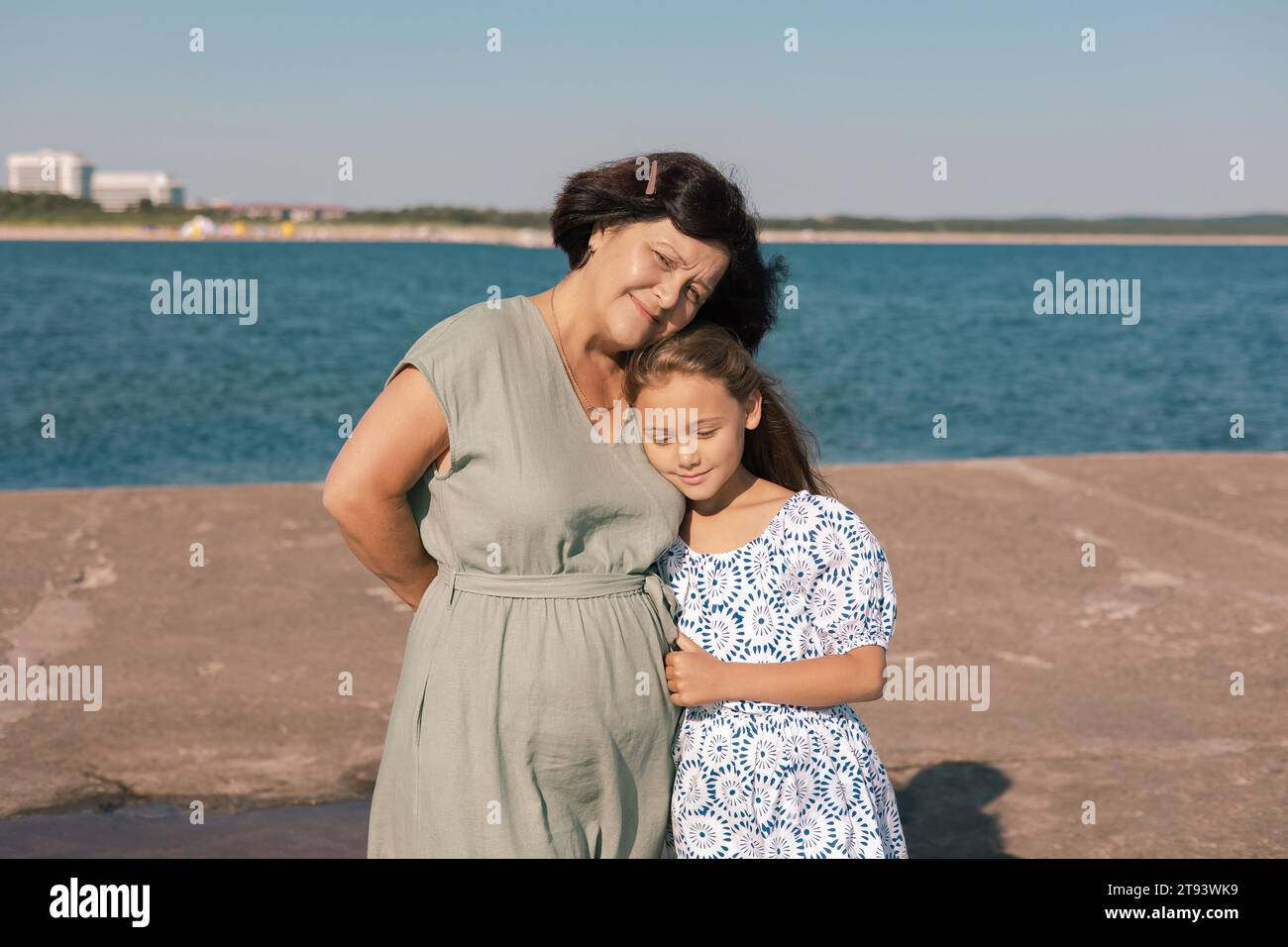 Una nonna con un abito verde abbraccia sua nipote con un abito a motivi sia in piedi in riva al mare con una costa lontana e edifici nel Foto Stock