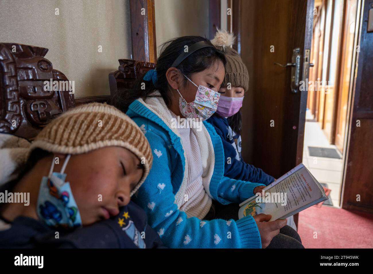 Viacha, la Paz, Bolivia – 16 agosto 2022: Tre bambini boliviani con maschere leggono un libro seduto su una panchina in una sala Foto Stock