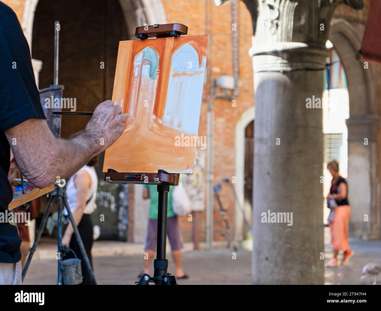 Primo piano della mano di un pittore mentre dipinge con pennello su tela una scena all'aperto in una piccola piazza a Venezia, Italia Foto Stock