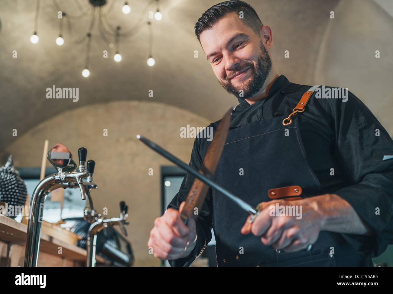 Cuoco barba sorridente con uniforme nera vestita con un coltello affilante a grembiule con l'utensile Honing Steel. Professione professionale, duro lavoro, cibo preparato Foto Stock