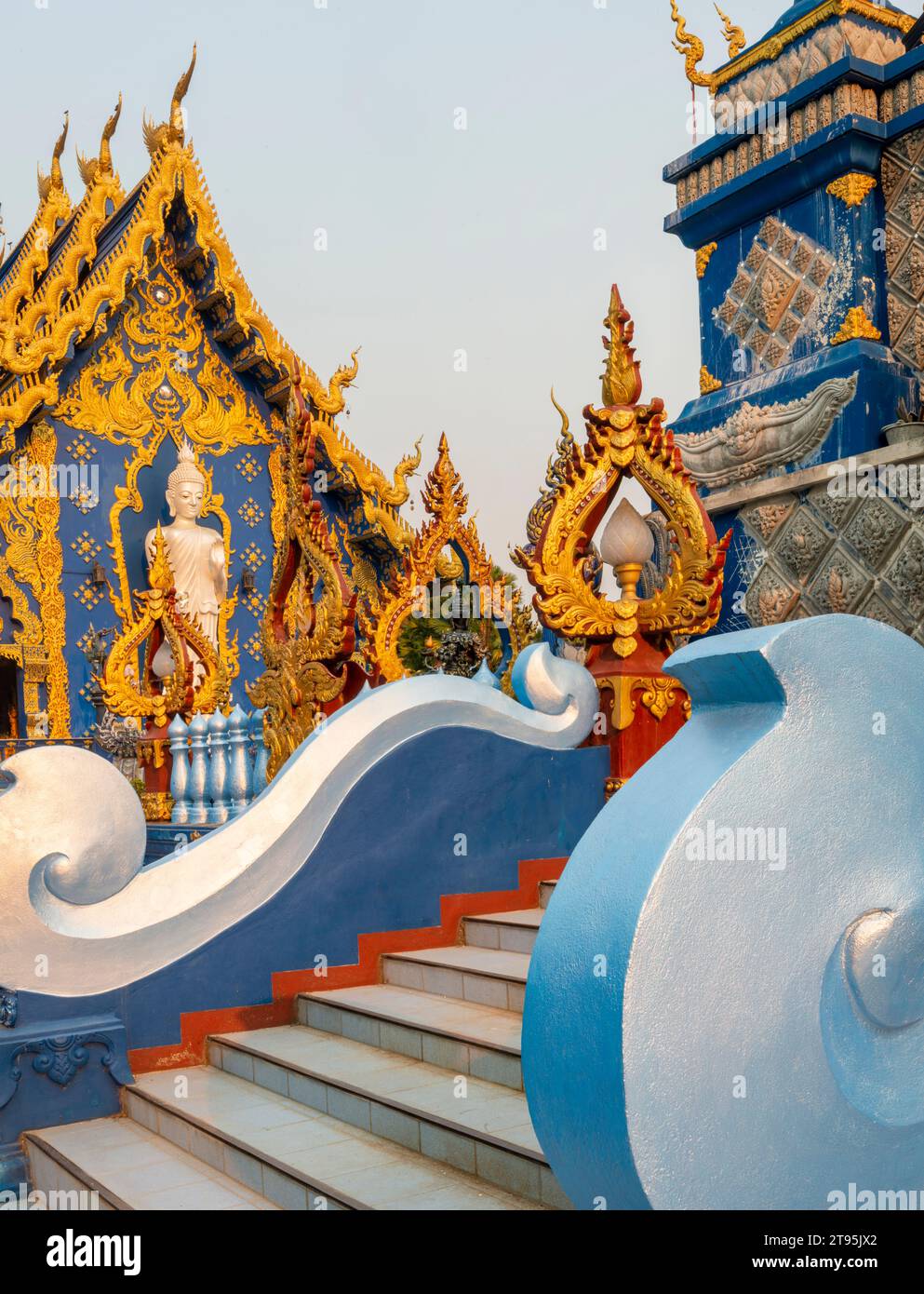 Esterno, splendidamente decorato con disegni blu e oro, statua di Buddha bianco-argento accanto all'ingresso del tempio e calda luce del tramonto che splende Foto Stock