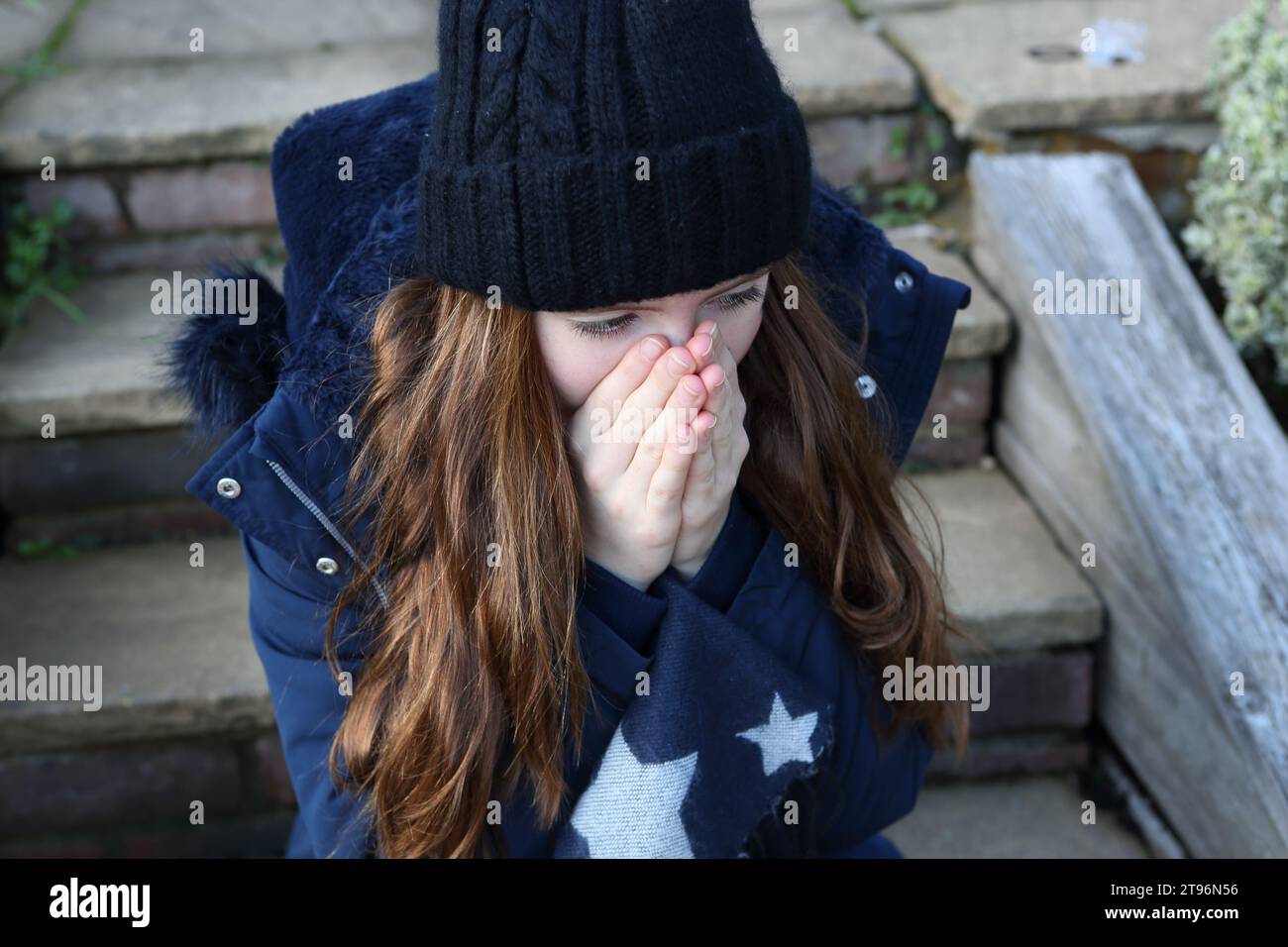 Ragazza adolescente che indossa un cappello di lana seduta sul gradino, con cappotto e sciarpa che cercano di tenersi al caldo Foto Stock