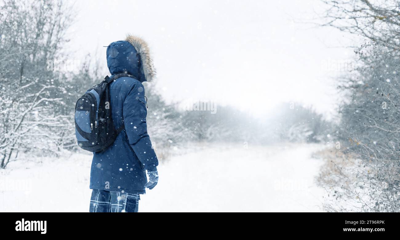 Vista posteriore del viaggiatore in una giacca blu con cappuccio di pelliccia e uno zaino sullo sfondo di un paesaggio invernale durante le nevicate Foto Stock