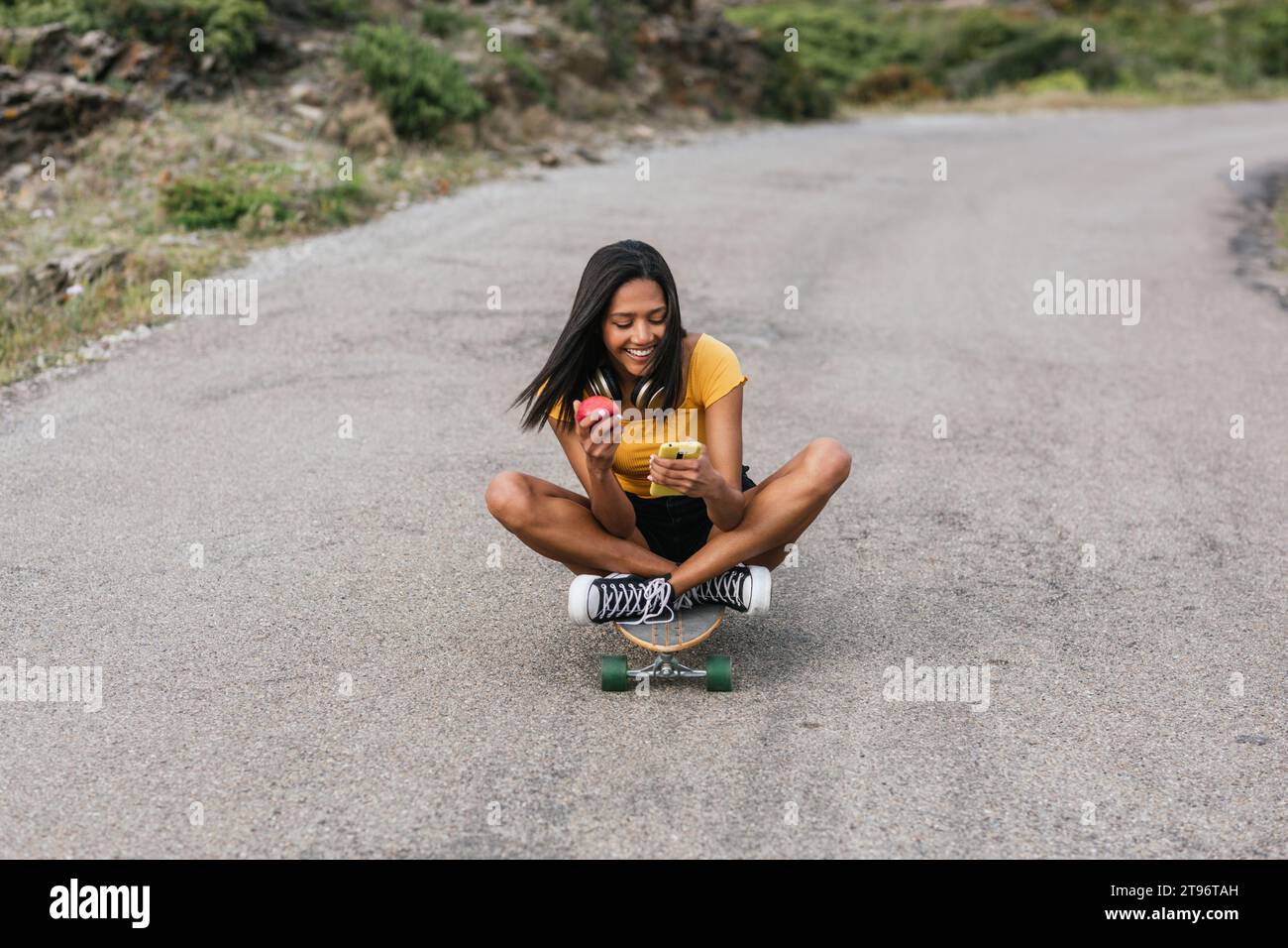 Corpo pieno di etnia positiva femmina con cuffie wireless sul collo e mela in mano guardando il cellulare mentre si guida longboard su strada asfaltata Foto Stock