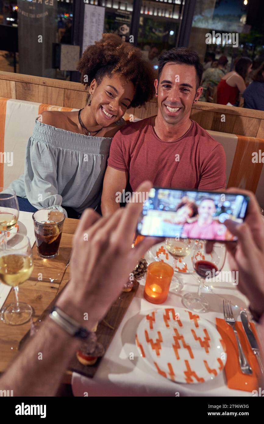 Dall'alto di uomini irriconoscibili che scattano foto di giovani coppie felici e diverse sedute al tavolo mentre cenano al pub Foto Stock