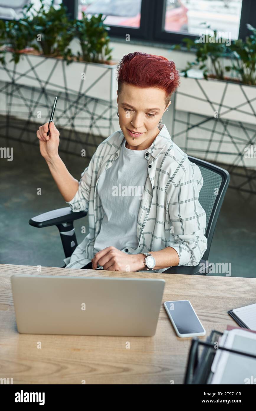 elegante persona non binaria con penna seduta sul posto di lavoro vicino a un computer portatile e a uno smartphone con schermo vuoto Foto Stock