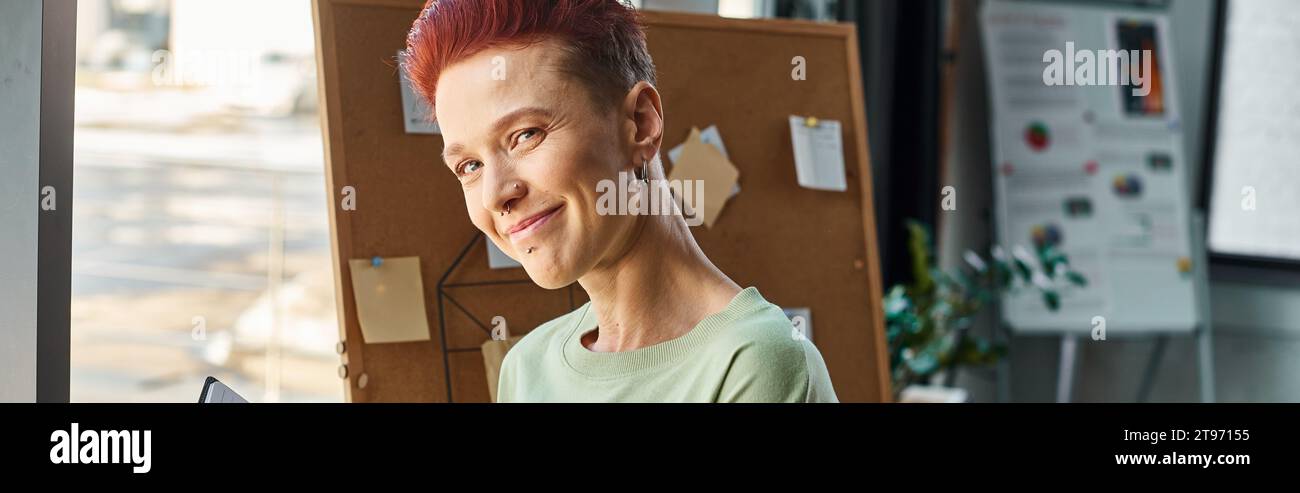 imprenditore non binario di successo che sorride davanti alla fotocamera in un moderno ambiente di ufficio, banner orizzontale Foto Stock