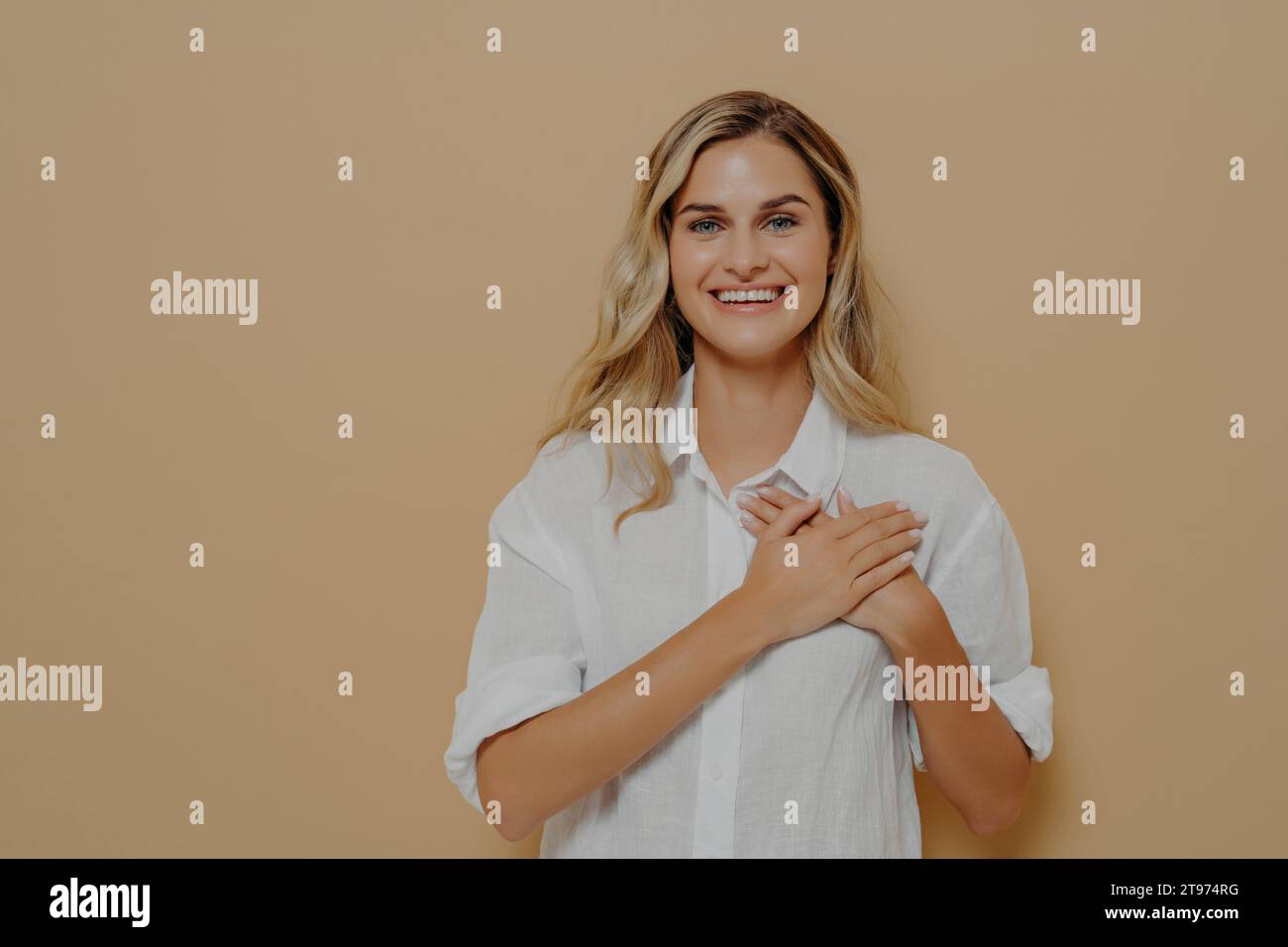 Donna radiosa in camicia bianca su beige, mani sul cuore, con un sorriso grato e luminoso Foto Stock