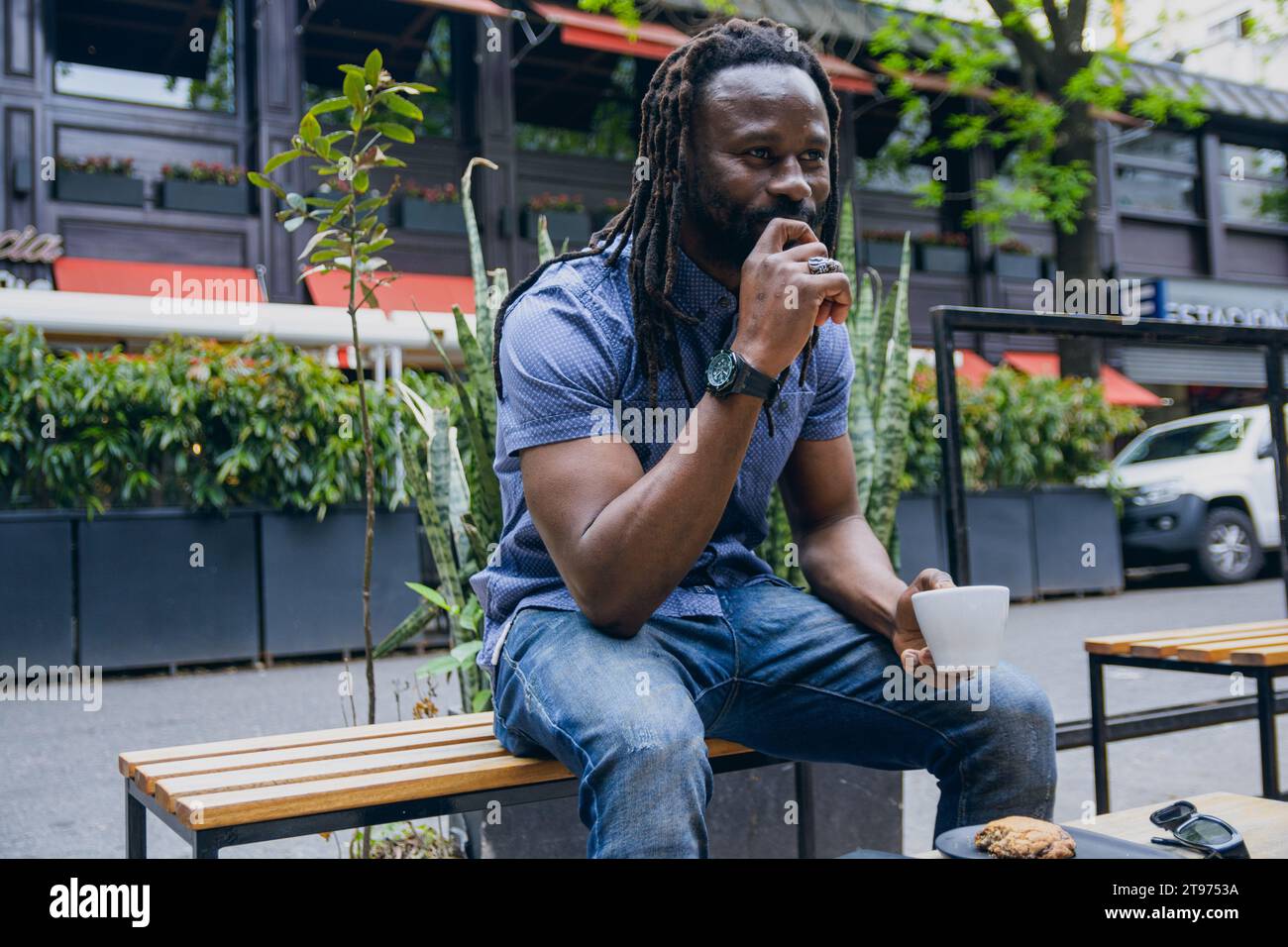 giovane africano con dreadlock seduti all'aperto a bere caffè e a pensare con la mano sulla bocca, guardando avanti meditando e godendosi il riposo Foto Stock