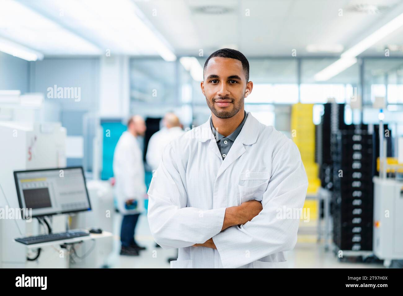 Tecnico sicuro di sé che sorride con le braccia incrociate mentre i colleghi parlano in una fabbrica di elettronica Foto Stock