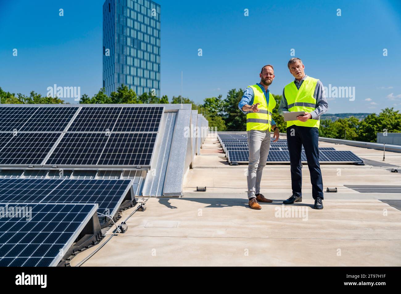 Due lavoratori in giubbotti di sicurezza discutono sul tetto di un edificio aziendale alimentato a energia solare mentre tengono in mano un tablet Foto Stock