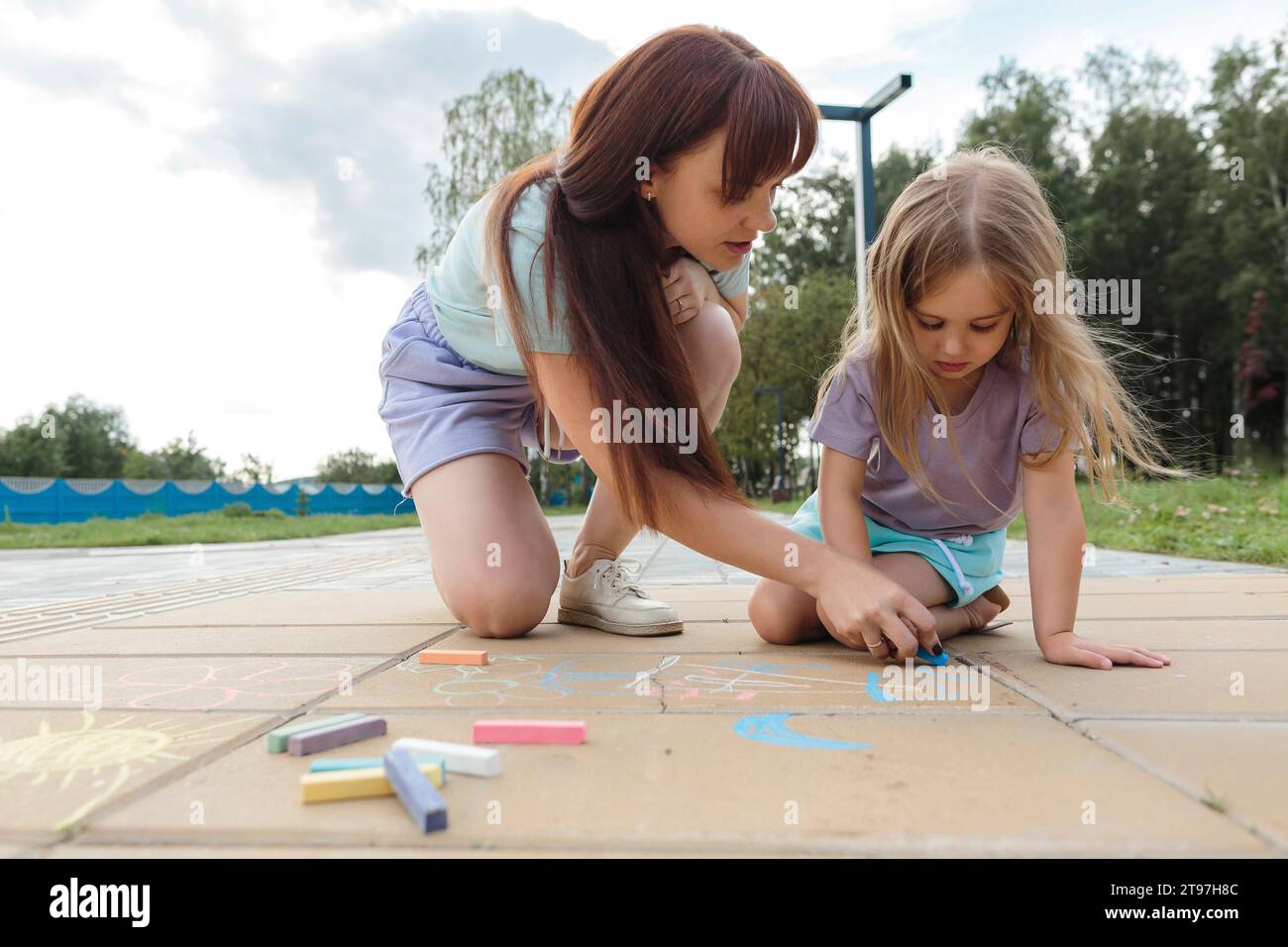 Donna che insegna a una figlia a disegnare con gesso sul pavimento piastrellato del parco Foto Stock