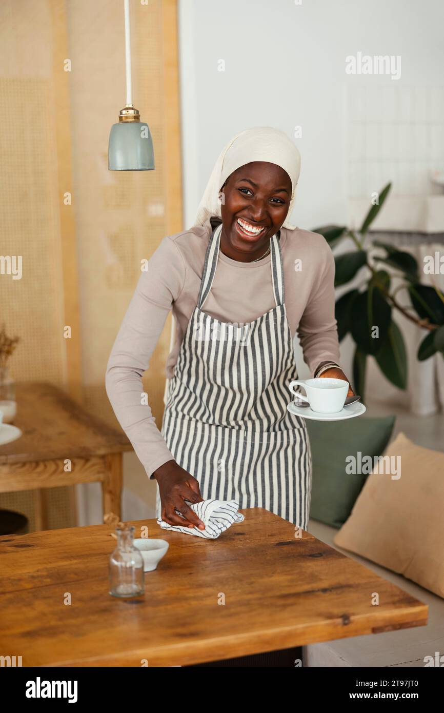 Barista sorridente che tiene una tazza di caffè vuota e un tavolo di pulizia nella caffetteria Foto Stock