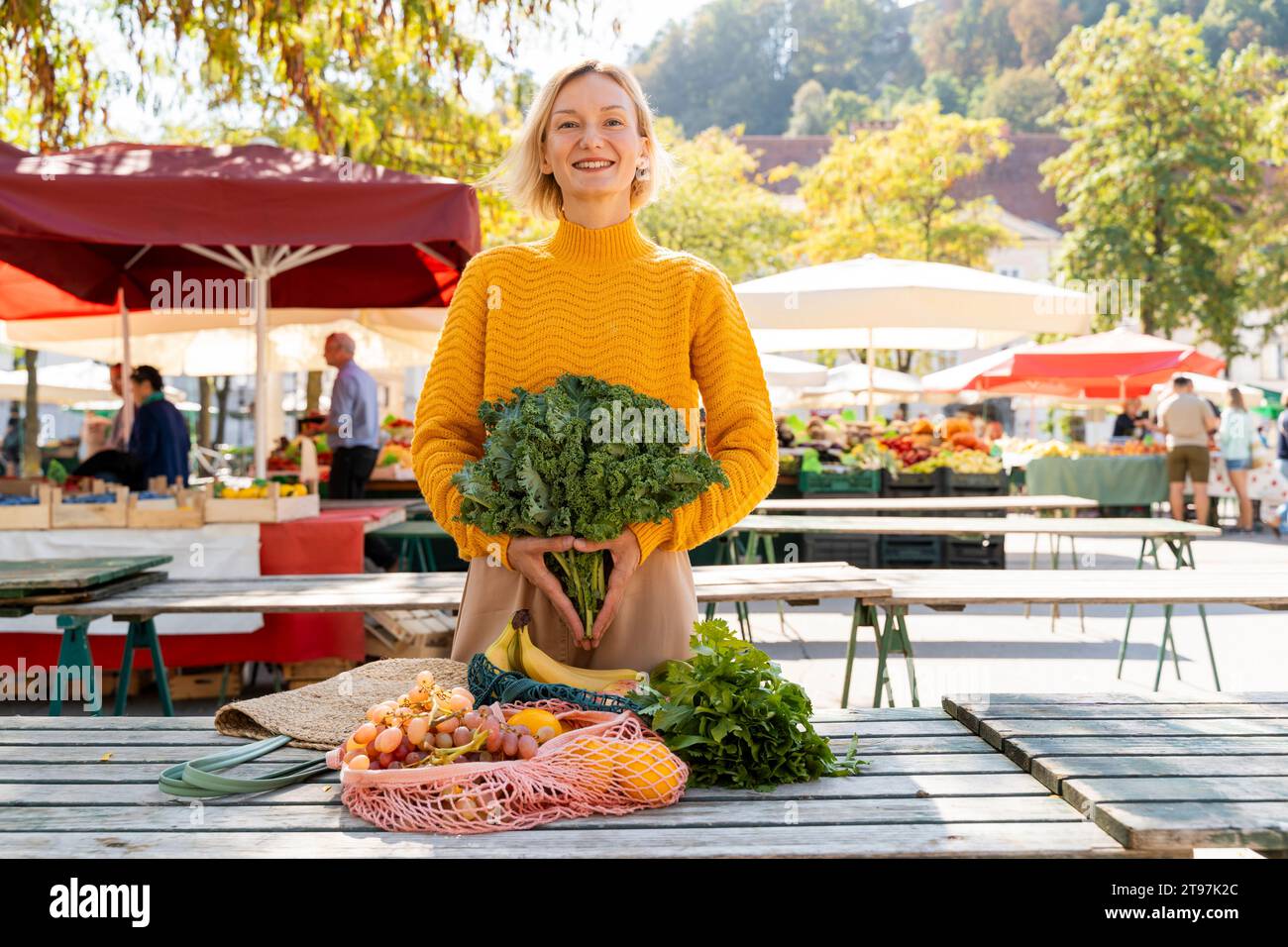 Donna sorridente che tiene verdure verdi a foglia vicino al tavolo al mercato agricolo Foto Stock