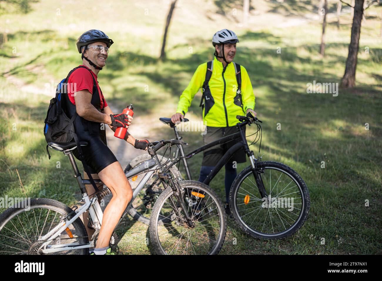 Uomo sorridente che tiene una bottiglia d'acqua e si trova vicino ad un amico con una bicicletta nella foresta Foto Stock
