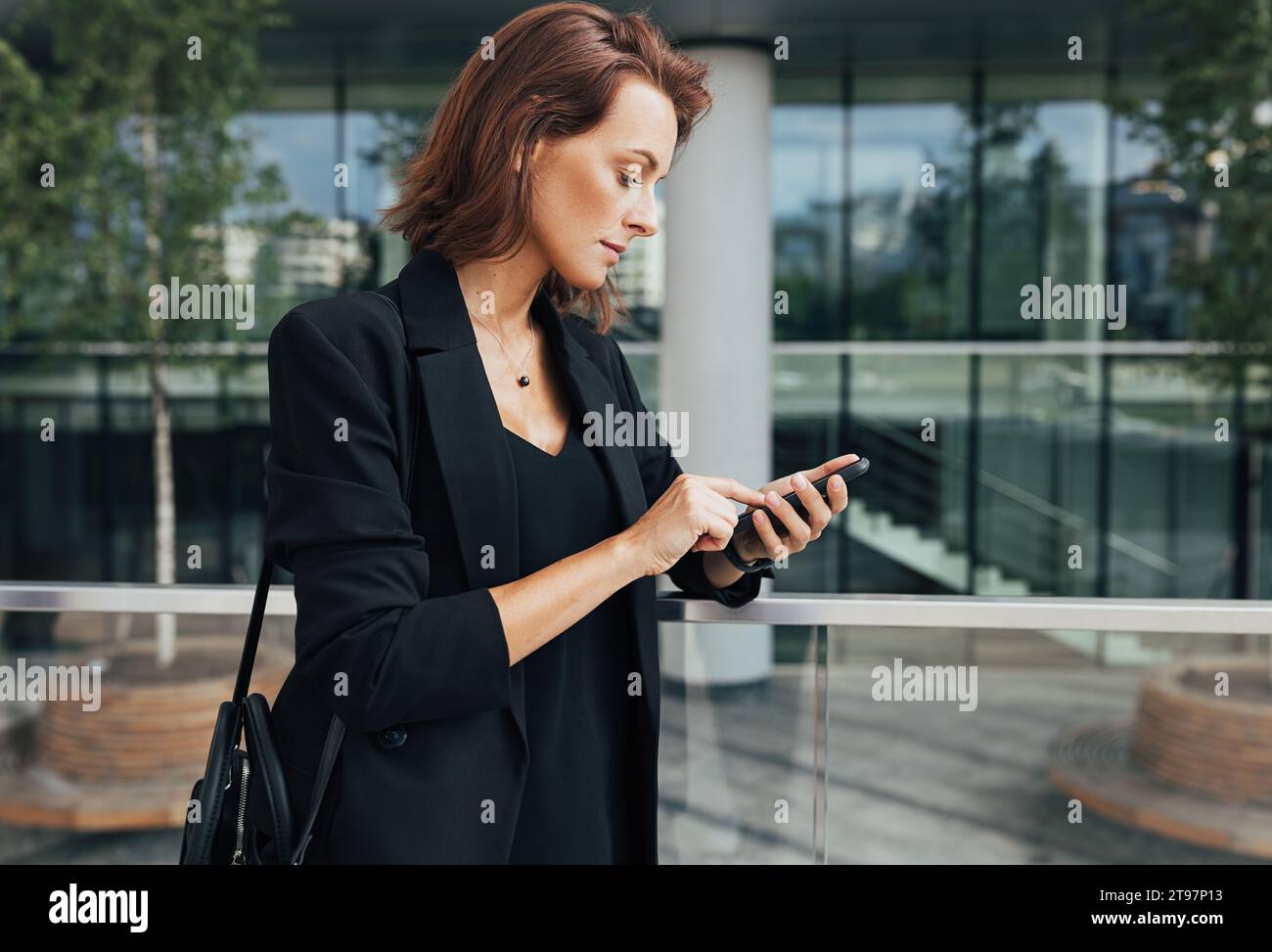 Vista laterale di una donna che indossa un abbigliamento formale che digita su uno smartphone. Donna di mezza età che tiene uno smartphone e sta in piedi all'aperto. Foto Stock