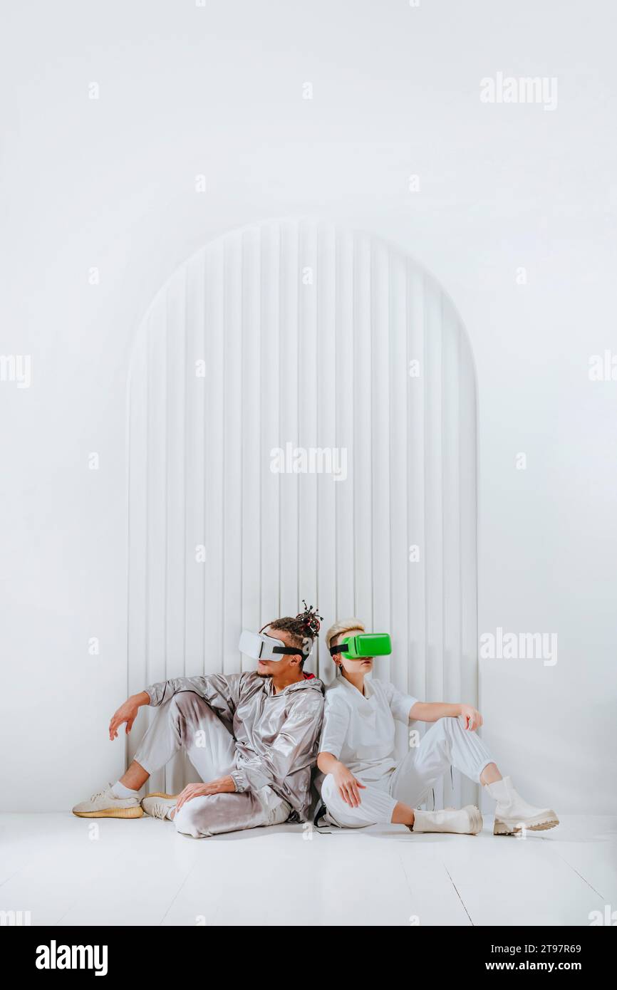 Giovane uomo e donna che indossa un visore per la realtà virtuale seduto dietro la schiena di fronte a una parete bianca Foto Stock