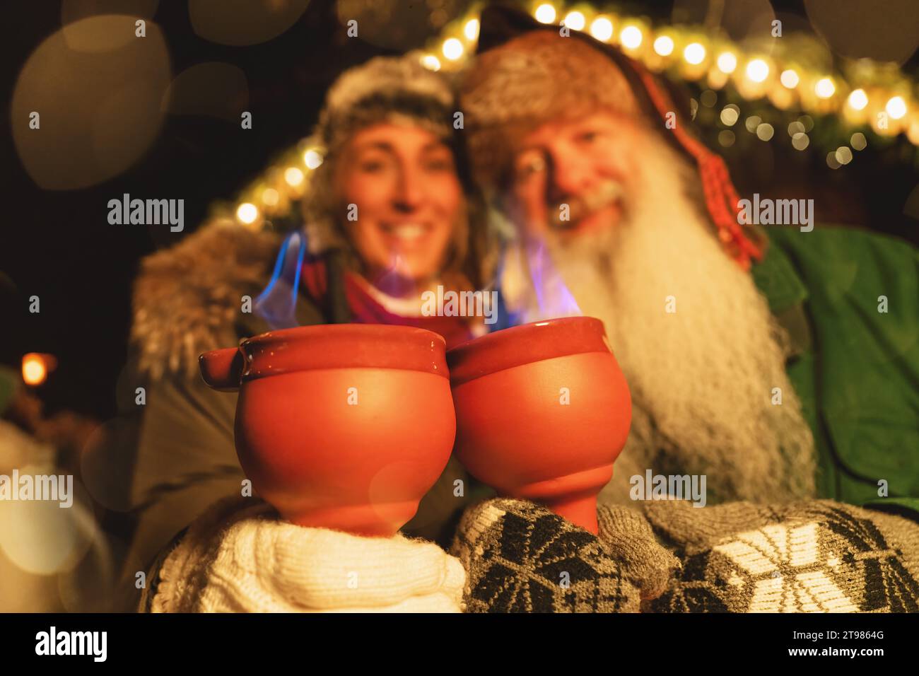 Gli amici brindano con le pinze fuochi, con fiamme blu sulle tazze, in un vivace mercatino di Natale Foto Stock