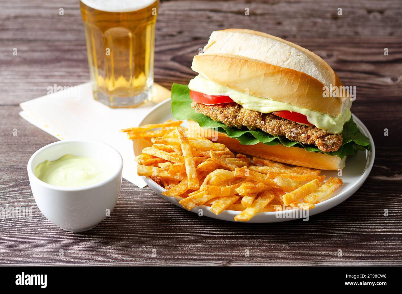 Panino alla milanesa con fette di pomodoro, lattuga e patatine fritte in un piatto bianco, una ciotola con maionese e un bicchiere di birra su fondo di legno. Foto Stock