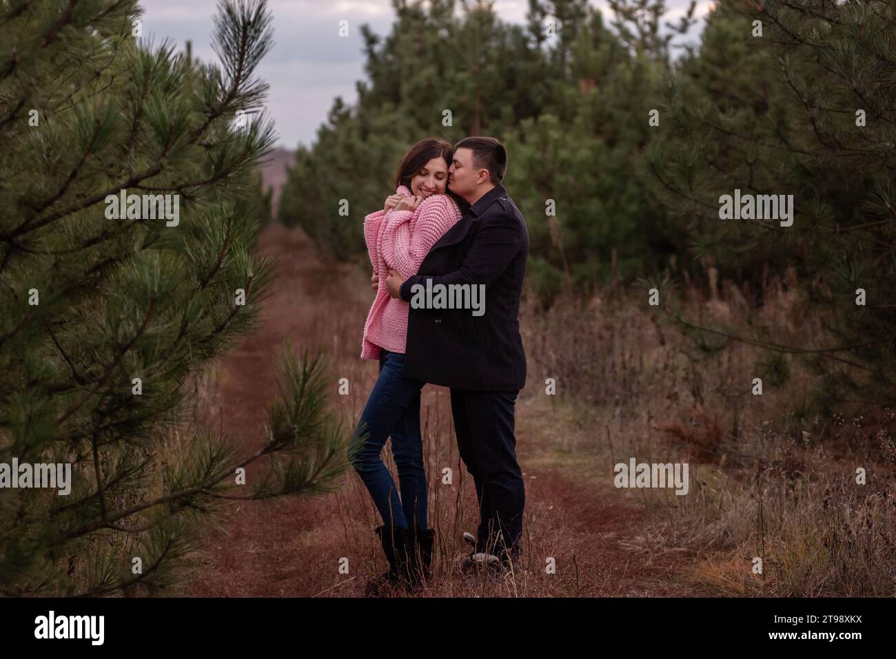 Ritratto ravvicinato della coppia felice che abbraccia la foresta di conifere verdi. Cappotto nero avvolto tattile intorno a una donna dai capelli ricci in un maglione rosa Foto Stock