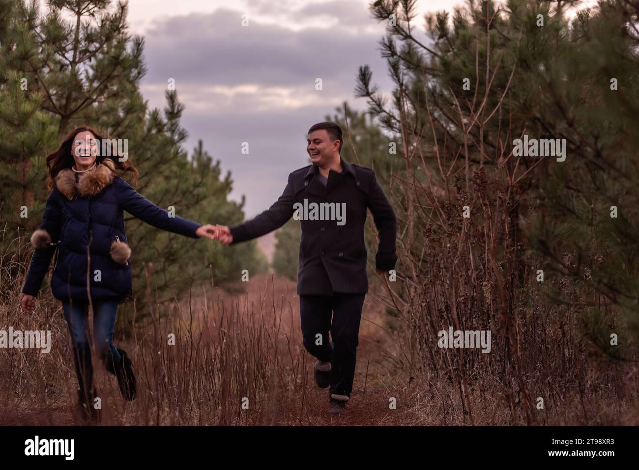 Coppia felice che corre attraverso la foresta tenendosi per mano. Un uomo con un cappotto nero e una giovane donna con un maglione a maglia si divertono in un parco di pini, pla Foto Stock