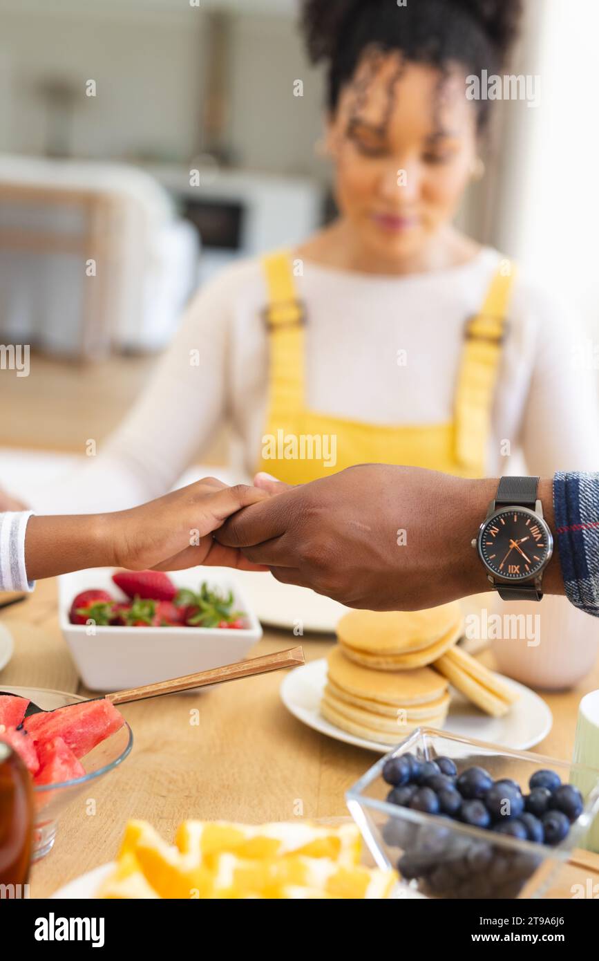 Famiglia afro-americana che si tiene per mano e prega al tavolo nella sala da pranzo a casa, spazio fotocopie. Insieme, genitorialità, infanzia, religione, cibo e.. Foto Stock