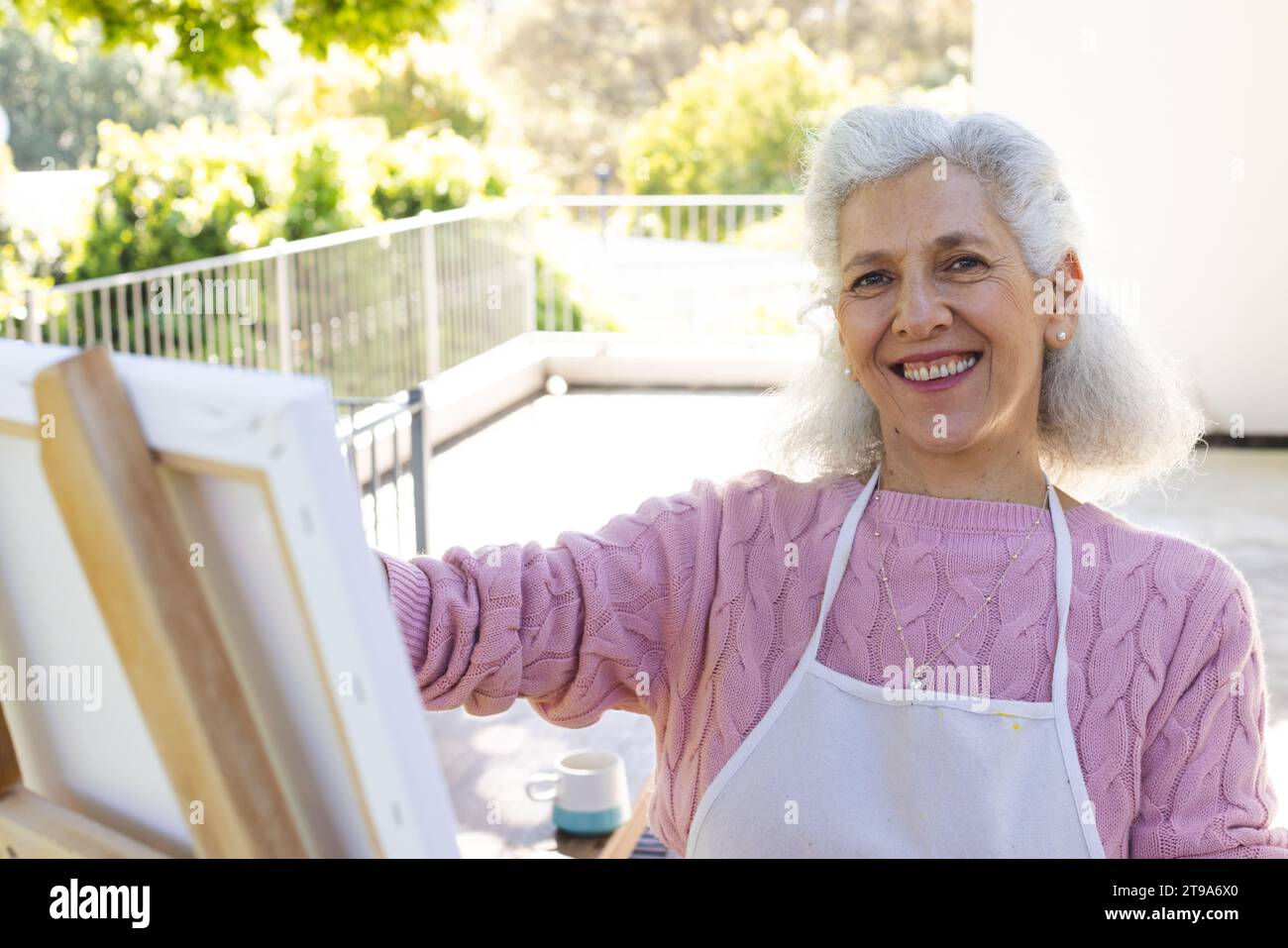 Ritratto di donna anziana caucasica felice che dipinge su tela su una terrazza soleggiata. Stile di vita, pensionamento, stile di vita senior, natura, creatività e domestico Foto Stock