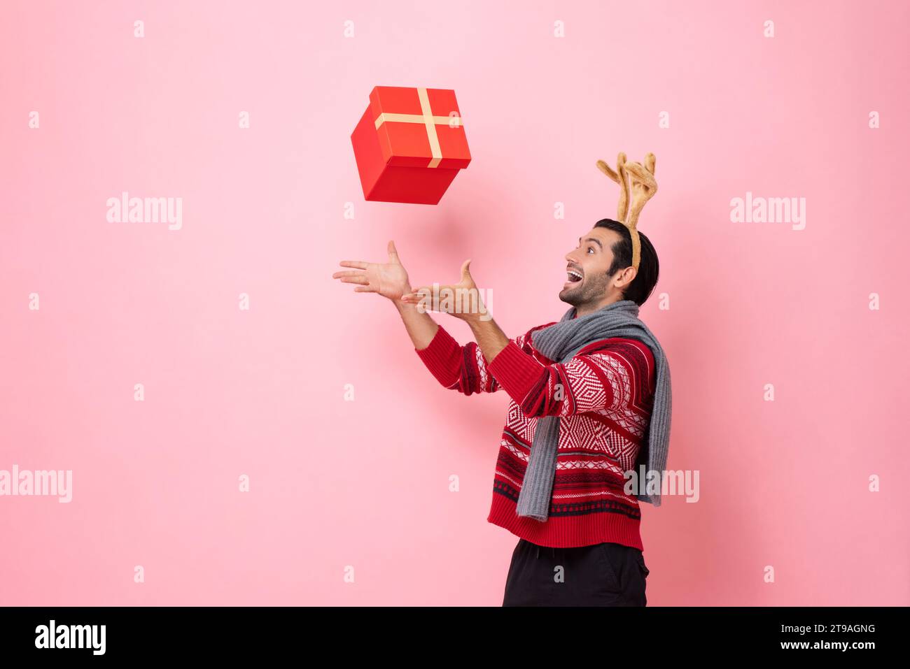 Ritratto in studio natalizio di un bell'uomo caucasico sorridente che indossa un maglione natalizio e una fascia da renna che riceve una scatola regalo in colore rosa iso Foto Stock