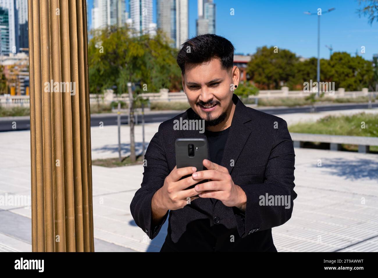 Primo piano di un giovane latinoamericano sorridente che controlla e risponde ai messaggi con il suo cellulare o smartphone in una piazza pubblica. Foto Stock